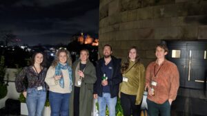 6 people stand casually next to each other smiling, it is night and edinburgh castle is lit up behind them