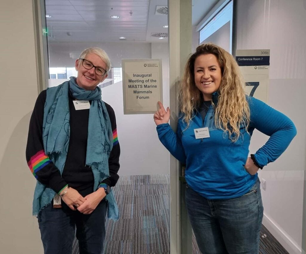 Two women standing in front of a meeting door and pointing to a sign saying Inaugural meeting of the marine mammals forum