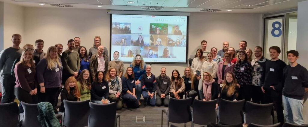 A group of people sitting and standing in a meeting room in front of a big screen smiling at the camera