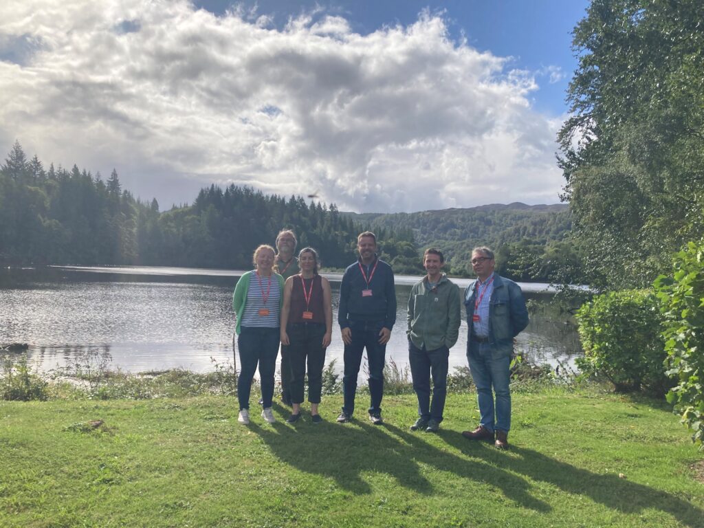 6 people standing by a lake, with a forest in the background